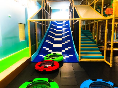 Colorful indoor play area featuring a blue slide, climbing structure, and inflatable rings on the floor.