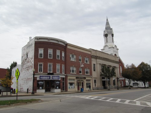 Historic buildings line a street, featuring a church steeple and a mix of brick and white architecture under a cloudy sky.