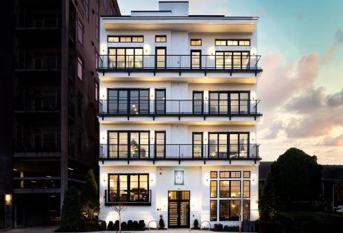 A modern, four-story white building with balconies, illuminated at dusk, surrounded by greenery and urban architecture.
