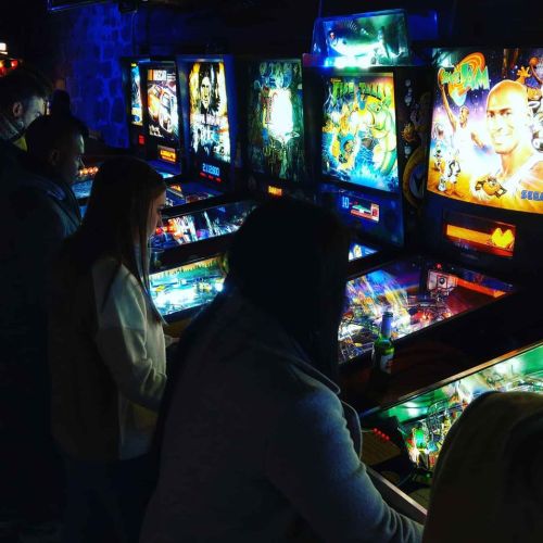 A dimly lit arcade with people playing colorful pinball machines. Bright lights reflect off the machines.