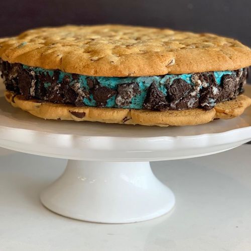 A large cookie sandwich filled with blue ice cream and chunks of chocolate cookies, displayed on a white cake stand.