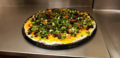 A large pizza topped with colorful vegetables, cheese, and olives on a black tray, set on a stainless steel surface.