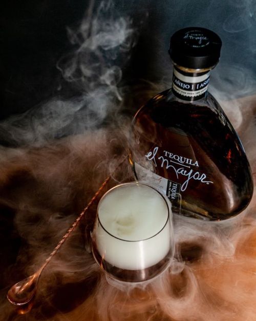 A bottle of El Mayor tequila beside a glass with a creamy drink, surrounded by swirling smoke.
