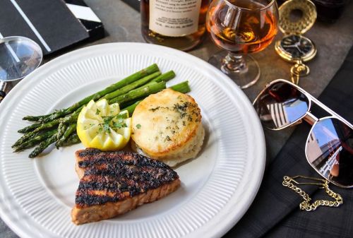 A plate with grilled salmon, asparagus, lemon, and a biscuit, accompanied by a glass of whiskey and stylish accessories.