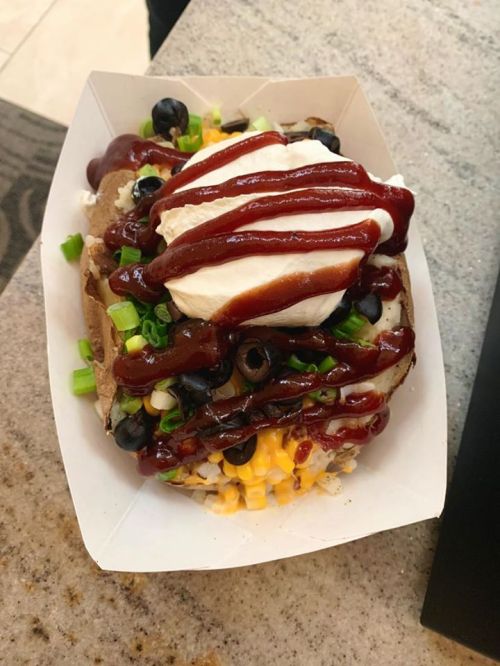 A loaded baked potato topped with sour cream, barbecue sauce, green onions, olives, corn, and blueberries in a paper tray.