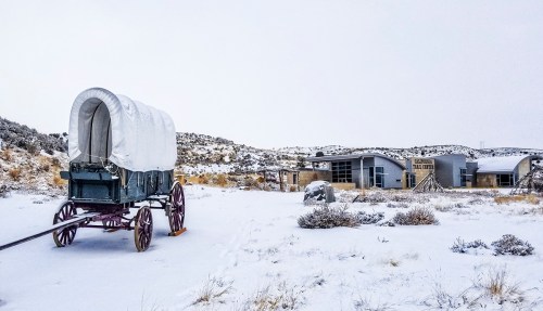 A covered wagon in a snowy landscape, with a modern building in the background and hills in the distance.