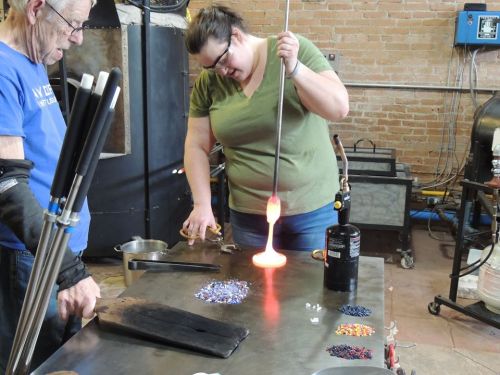 A woman shapes molten glass with a blowpipe while a man observes in a glassblowing studio. Colorful beads are scattered on the table.