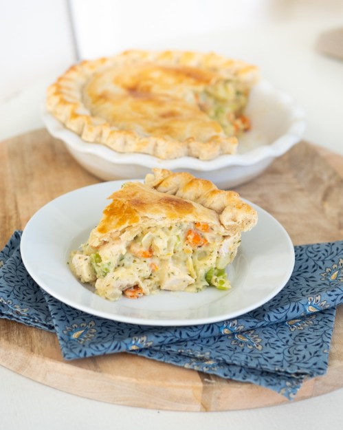 A slice of chicken pot pie on a plate, with the whole pie in a dish behind it, on a wooden cutting board.
