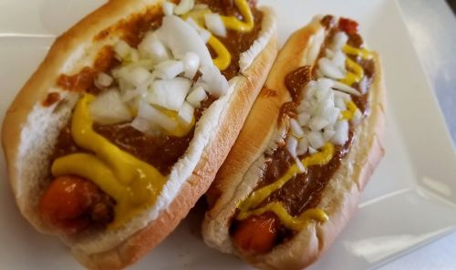 Two chili cheese hot dogs topped with mustard and diced onions, served on a white plate.