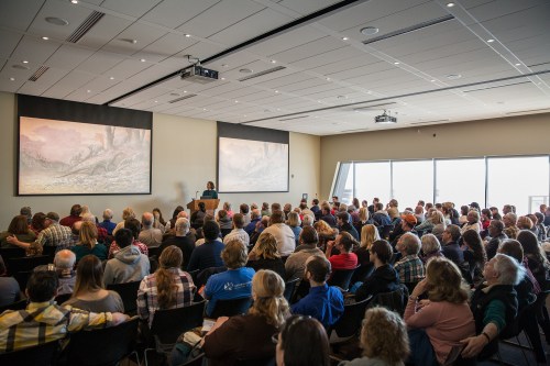 A large audience listens attentively to a speaker in a modern conference room with large screens.