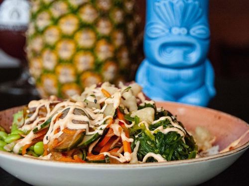 A colorful plate of vegetables drizzled with sauce, with a blue tiki statue and a pineapple in the background.