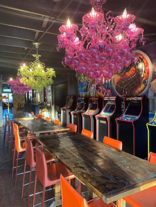 A vibrant arcade with vintage game machines, a long wooden table, and colorful chandeliers hanging above.