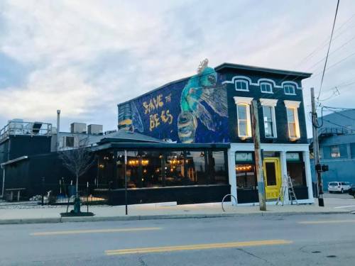 A colorful mural reading "Save the Bees" on the side of a stylish restaurant building at dusk.