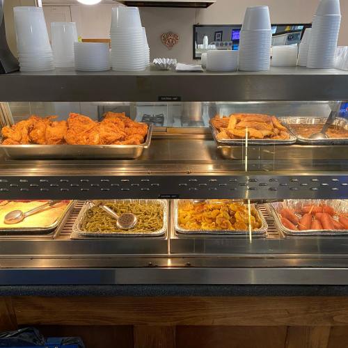 A buffet display featuring fried chicken, fries, green beans, and sliced hot dogs in metal trays.