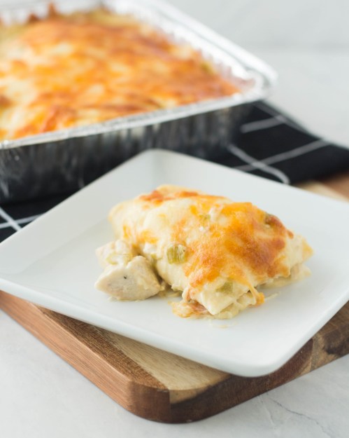 A serving of cheesy chicken enchiladas on a white plate, with a baking dish of enchiladas in the background.