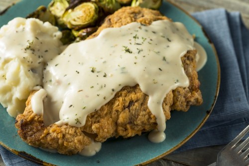 A plate of fried chicken topped with creamy gravy, served with mashed potatoes and roasted Brussels sprouts.
