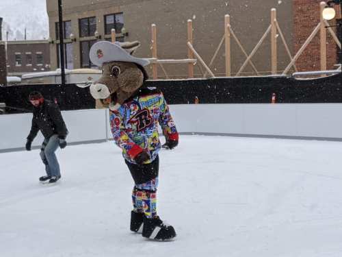 A colorful mascot in a winter outfit skating on an ice rink while snow falls around.