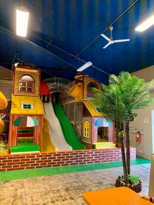 Indoor play area featuring a colorful slide, playhouse structures, and a decorative palm tree.