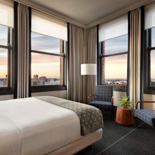 Modern hotel room with a large bed, stylish chairs, and windows showcasing a sunset view.