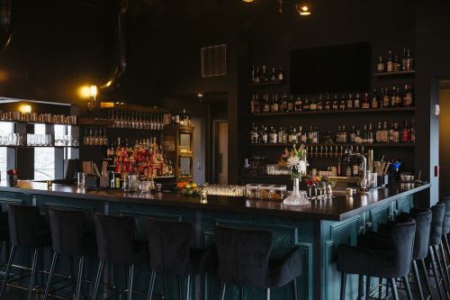A stylish bar with a dark interior, featuring a well-stocked shelf of spirits and a sleek counter with high stools.