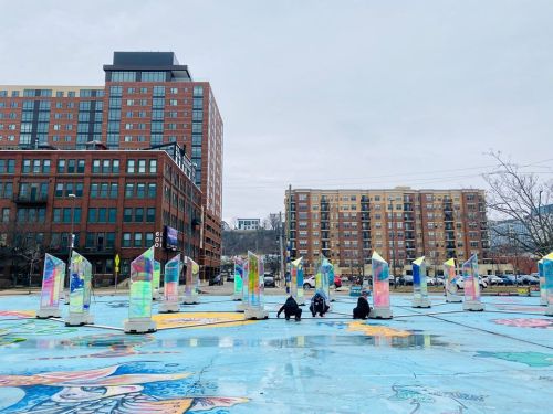 A colorful outdoor art installation with mirrored structures and people sitting on a painted surface, surrounded by buildings.