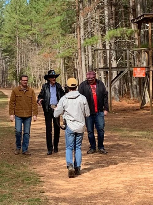 A group of four men walking on a wooded path, surrounded by tall trees and a wooden structure in the background.