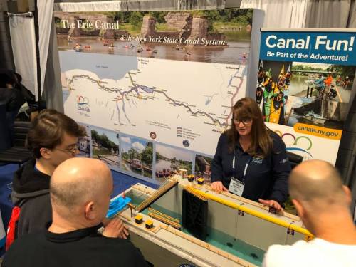 A woman explains the Erie Canal model to a small group at a booth featuring the New York State Canal System.