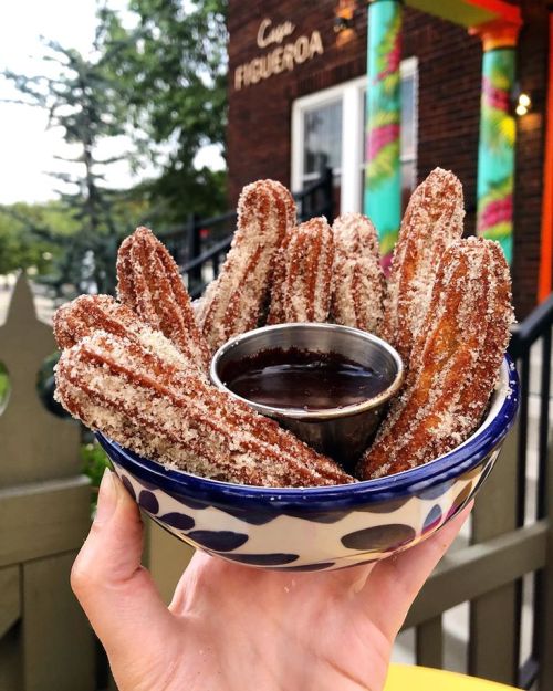 A hand holding a bowl of churros coated in sugar, with a small cup of chocolate sauce for dipping.