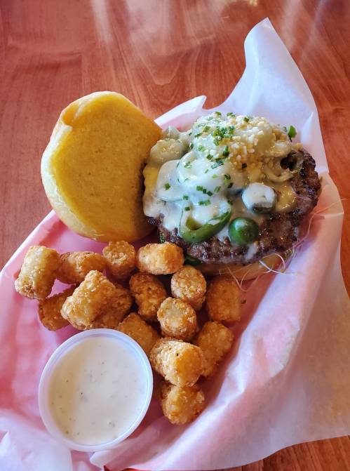 A plate featuring a cheesy burger with jalapeños, a side of tater tots, and a piece of cornbread.
