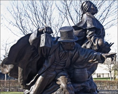 A bronze statue depicting a man with a lantern and a woman, both in historical attire, set against a bare tree backdrop.