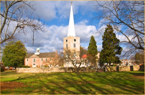 A historic church with a tall white steeple, surrounded by trees and a grassy area under a partly cloudy sky.
