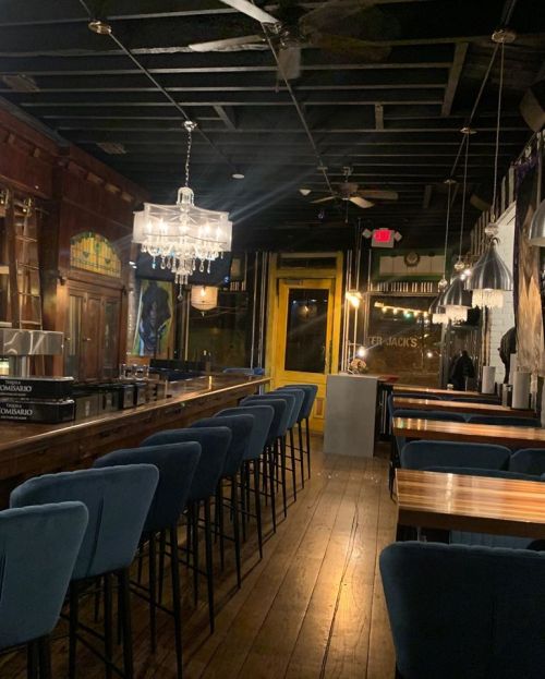 A dimly lit restaurant interior featuring a long bar, blue chairs, wooden tables, and a chandelier.
