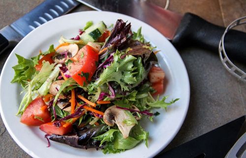 A colorful salad with mixed greens, tomatoes, cucumbers, mushrooms, and carrots on a white plate, surrounded by kitchen tools.