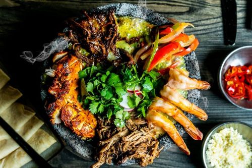 A vibrant platter of grilled meats, shrimp, peppers, and fresh herbs, served in a stone bowl with tortillas on the side.