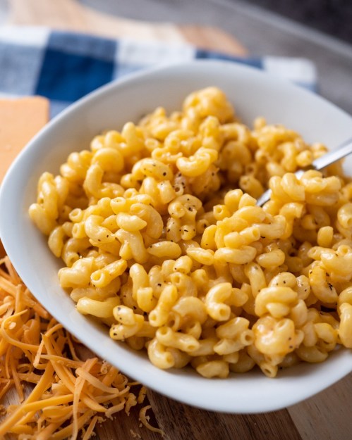 A bowl of creamy macaroni and cheese with a fork, surrounded by shredded cheese on a wooden surface.