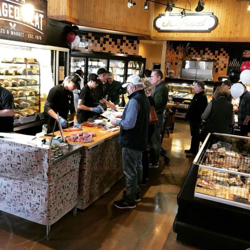 A busy market scene with customers lined up at a meat counter, interacting with staff and browsing products.