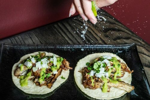 A hand squeezes lime over two tacos filled with meat, onions, and green sauce on a black plate.