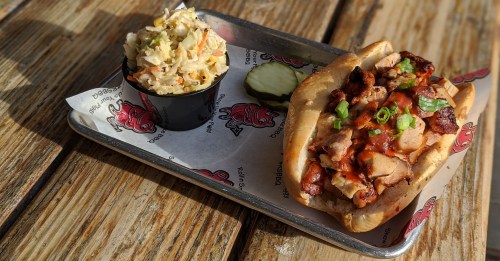 A tray with a BBQ chicken sandwich topped with green onions, a side of coleslaw, and a pickle slice on a wooden table.