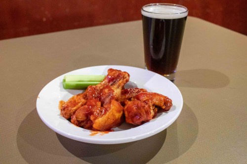 A plate of saucy chicken wings with a celery stick, accompanied by a glass of dark beer on a table.