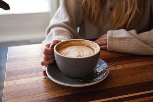 A person holds a large cup of latte art on a wooden table, with a spoon resting on the saucer.