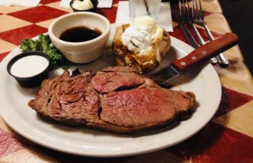 A plate featuring a juicy slice of prime rib, a baked potato with sour cream, and dipping sauces on a checkered tablecloth.