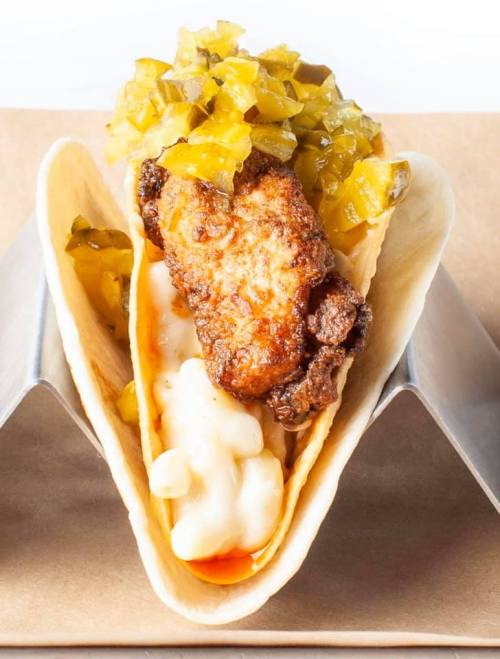 A taco filled with fried chicken, creamy sauce, and topped with pickles and diced green peppers, served in a taco holder.
