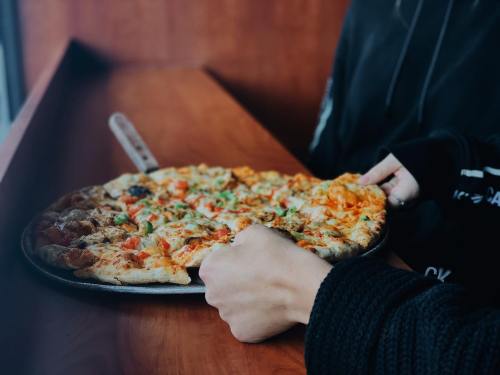 A person holding a pizza on a wooden table, with various toppings visible.
