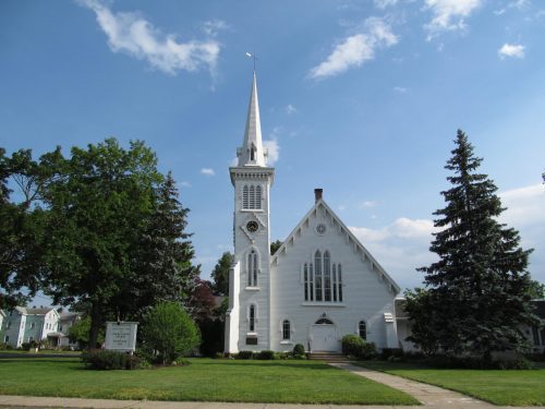 A white church with a tall steeple, surrounded by green trees and a clear blue sky.
