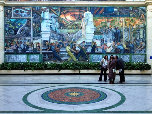 A large mural depicting industrial workers and machinery, with people standing in front of it in a spacious hall.