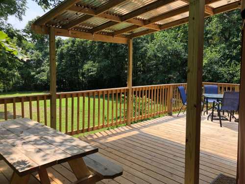 A wooden deck with a picnic table and chairs, surrounded by greenery and trees in a sunny outdoor setting.