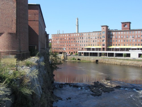 A historic mill building beside a calm river, with a clear blue sky and industrial structures in the background.