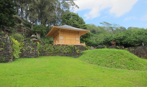 A serene wooden structure sits atop a grassy hill, surrounded by lush greenery and stone walls under a blue sky.