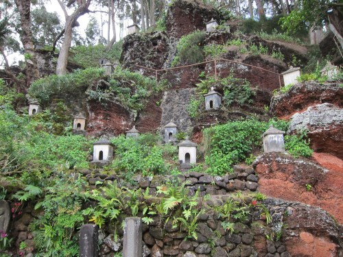 A hillside covered in greenery and small stone structures, surrounded by trees and rocky terrain.
