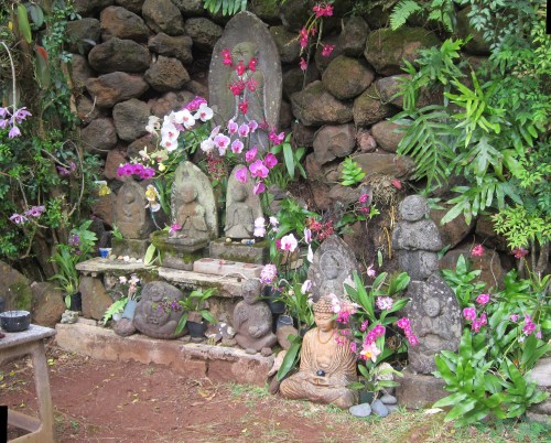 A serene garden scene featuring stone statues surrounded by vibrant orchids and lush greenery against a rocky backdrop.